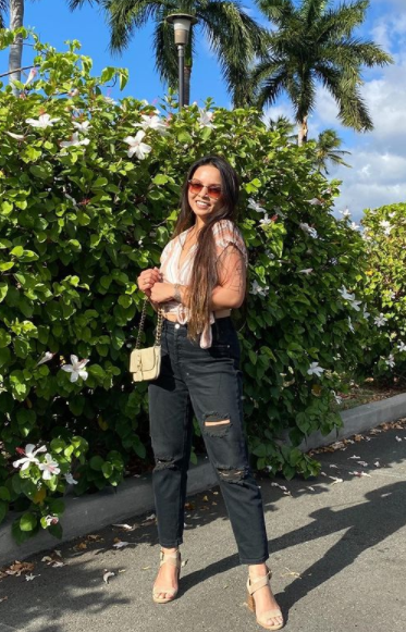 Girl smiling at the camera. Wearing striped crop blouse and black distressed jeans with neutral block heels and holding chained cream shoulder bag